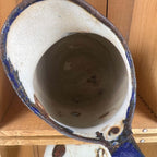 Overhead close-up of the opening and interior of a white enamelware pitcher. Brown staining and scattered coating losses appear inside, while chipped cobalt-blue enamel and exposed dark metal surround the rim. Part of the handle is visible at the lower right.