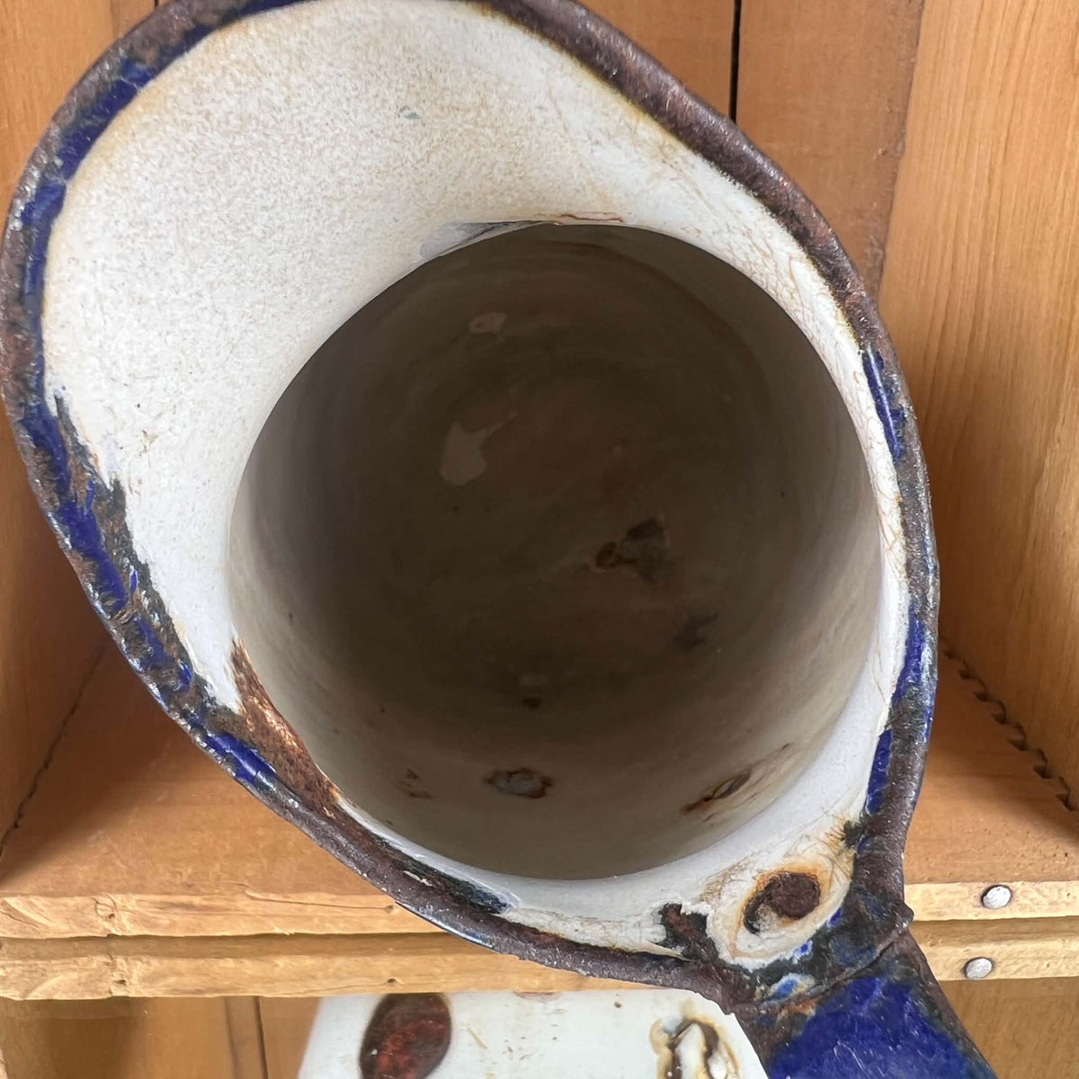 Overhead close-up of the opening and interior of a white enamelware pitcher. Brown staining and scattered coating losses appear inside, while chipped cobalt-blue enamel and exposed dark metal surround the rim. Part of the handle is visible at the lower right.