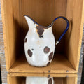 White enamelware duckbill jug standing upright inside a wooden crate, with an elongated pouring spout on the left and curved cobalt-blue handle on the right. Large brown patches of exposed metal interrupt the rounded body, with chipped coating along the upper rim.