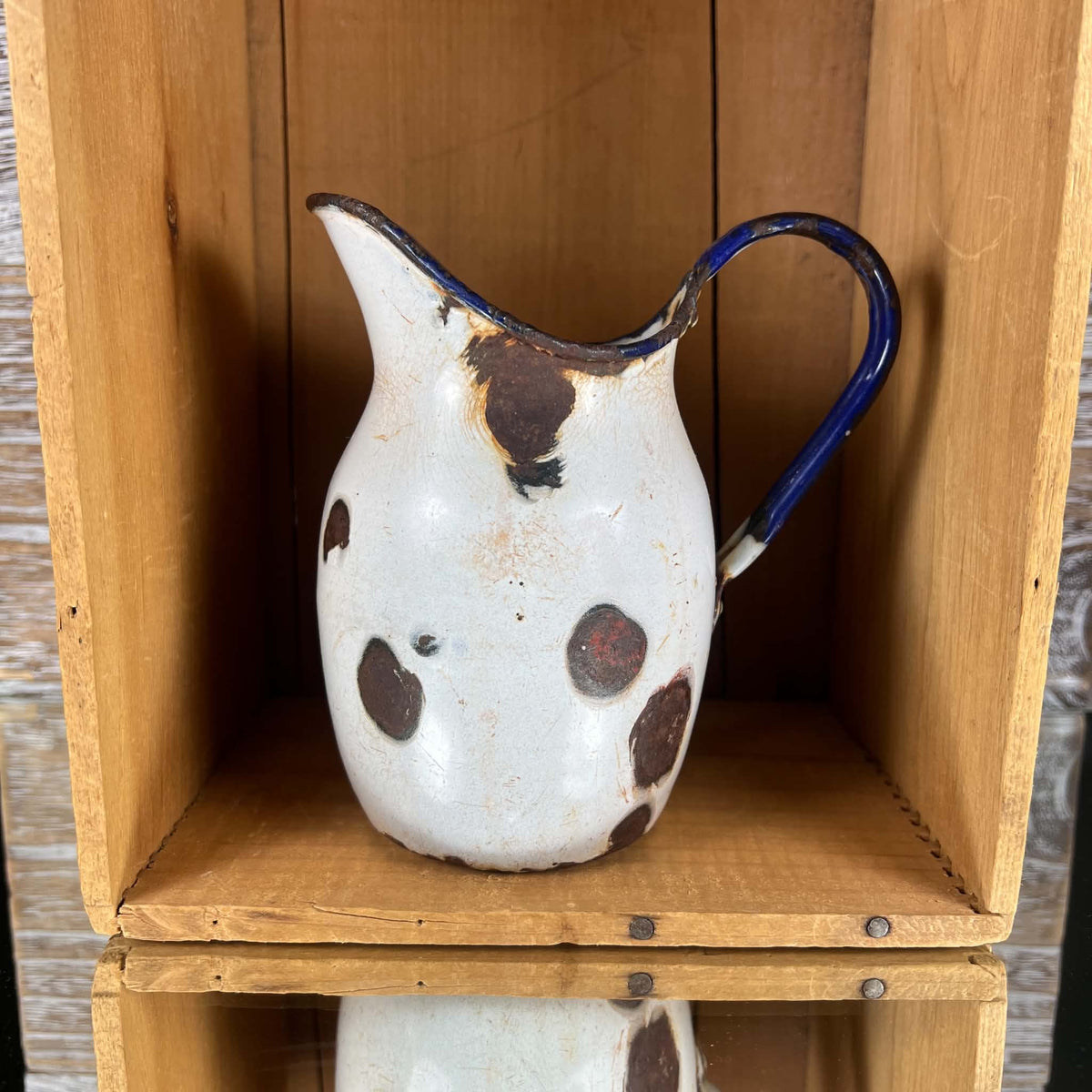White enamelware duckbill jug standing upright inside a wooden crate, with an elongated pouring spout on the left and curved cobalt-blue handle on the right. Large brown patches of exposed metal interrupt the rounded body, with chipped coating along the upper rim.