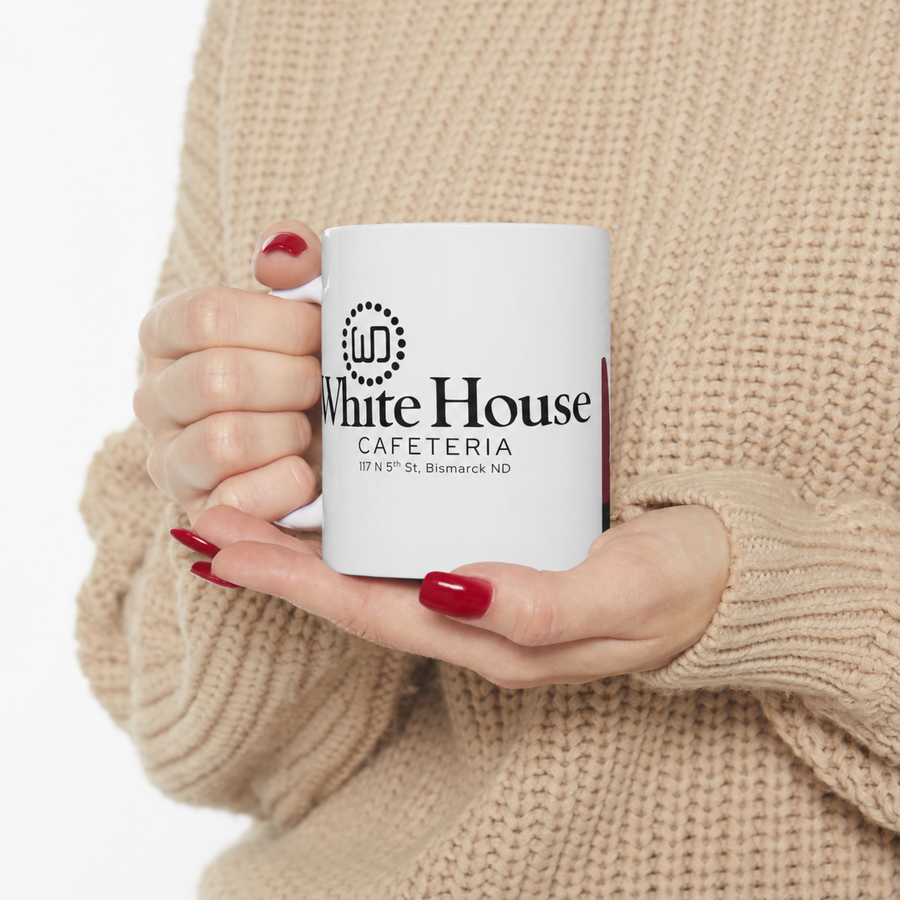 White House Cafeteria ceramic coffee mug held in both hands against a chunky tan knit sweater with glossy red nails. The mug displays the original cafe logo and 117 N 5th St, Bismarck, ND address, creating a warm lifestyle view of this nostalgic local keepsake.