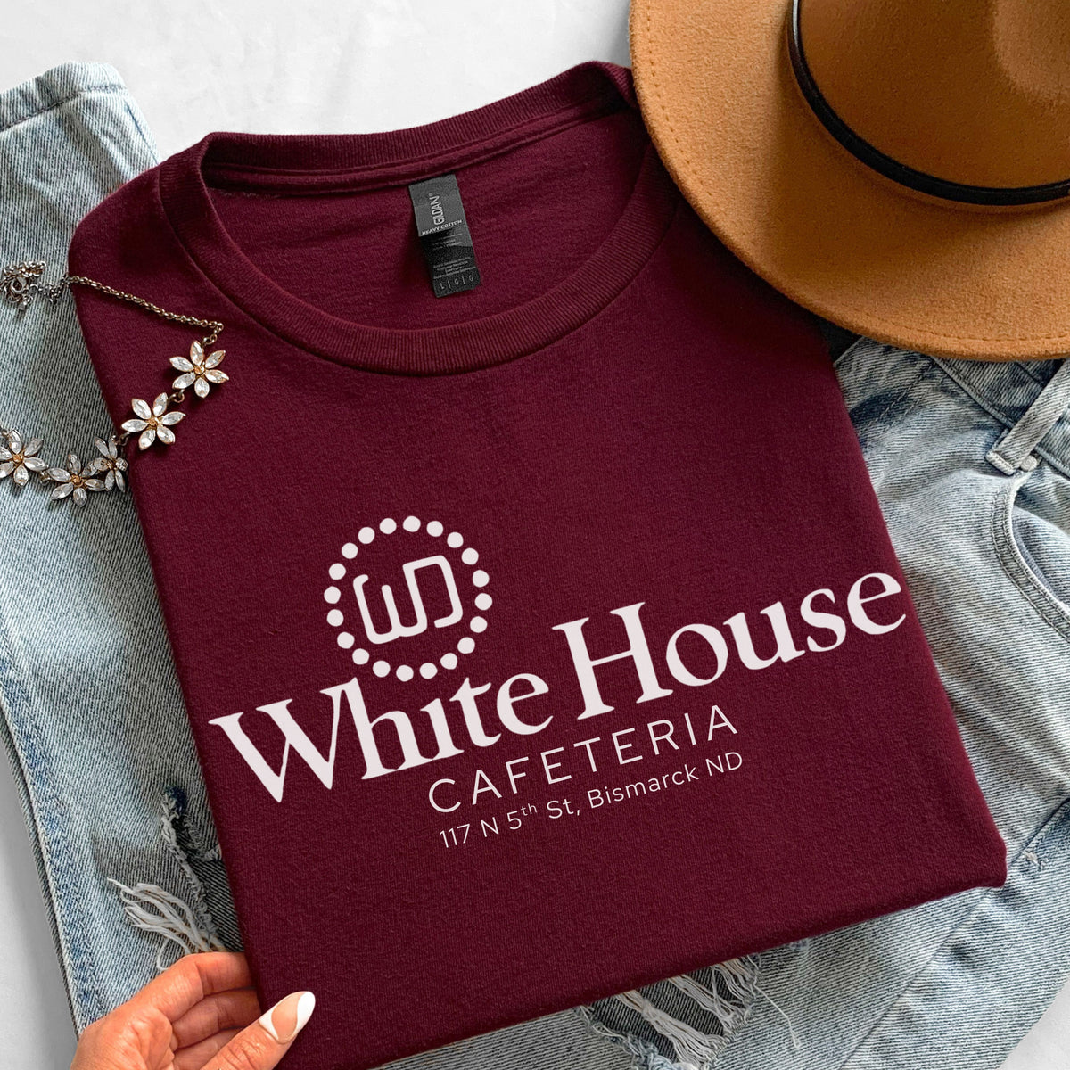 Maroon White House Cafeteria T-shirt arranged in a casual flatlay with denim, a tan felt hat and small floral accents. The front displays the original white cafe logo and 117 North 5th Street address, commemorating the longtime Bismarck, North Dakota restaurant.
