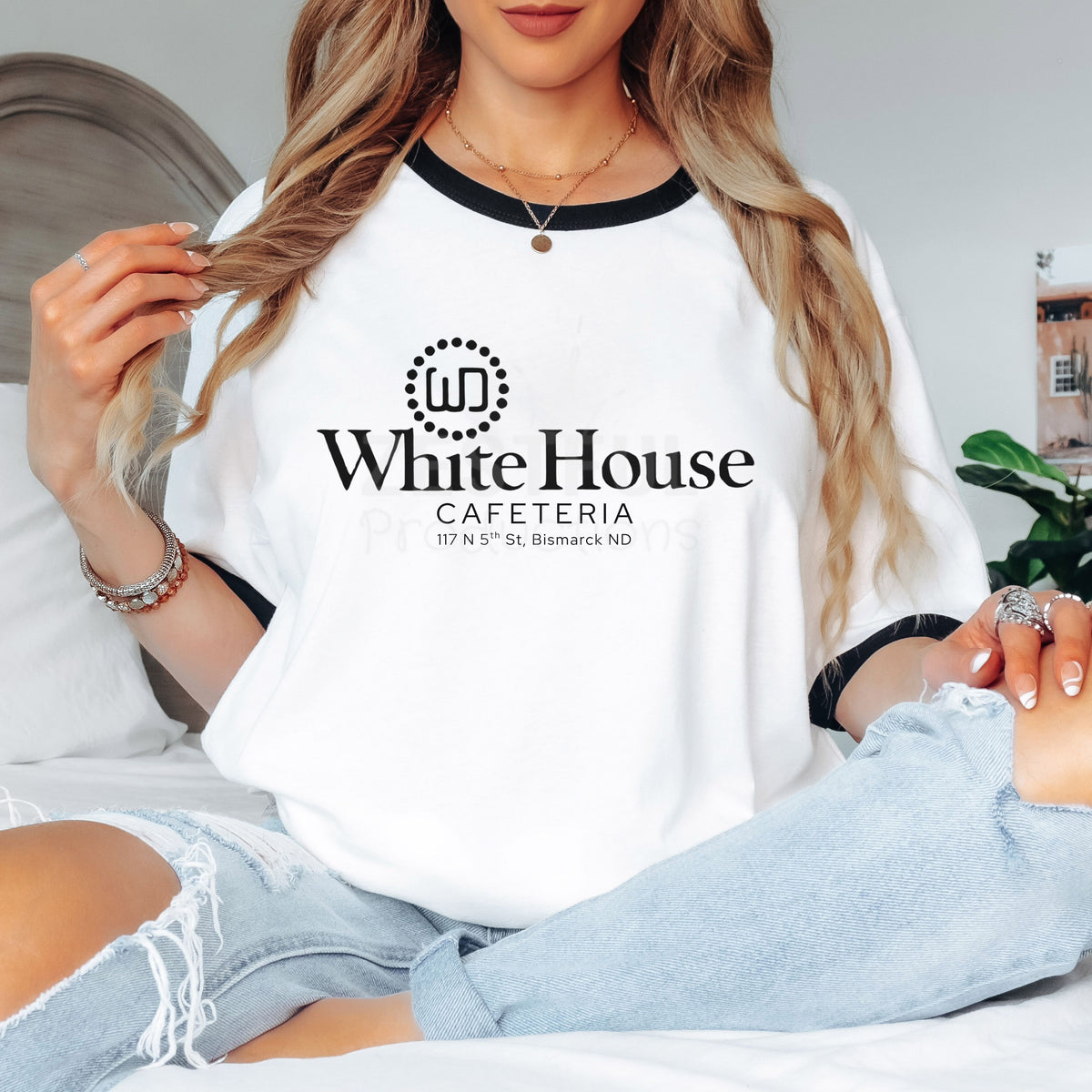 Woman seated casually in light-wash jeans wearing a white retro ringer T-shirt with black neckline and sleeve bands. The original White House Cafeteria logo and 117 North 5th Street address appear on the front, honoring the longtime Bismarck, North Dakota restaurant.