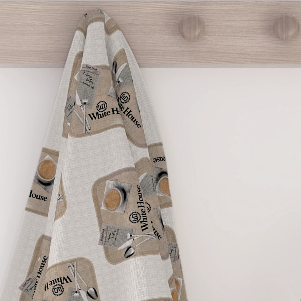 White House Cafeteria kitchen tea towel hanging from a light wood wall hook. The soft, light oatmeal towel displays a repeating pattern of vintage cafeteria trays with coffee, silverware, a handwritten Cheese Buttons and caramel roll order, and the White House logo.