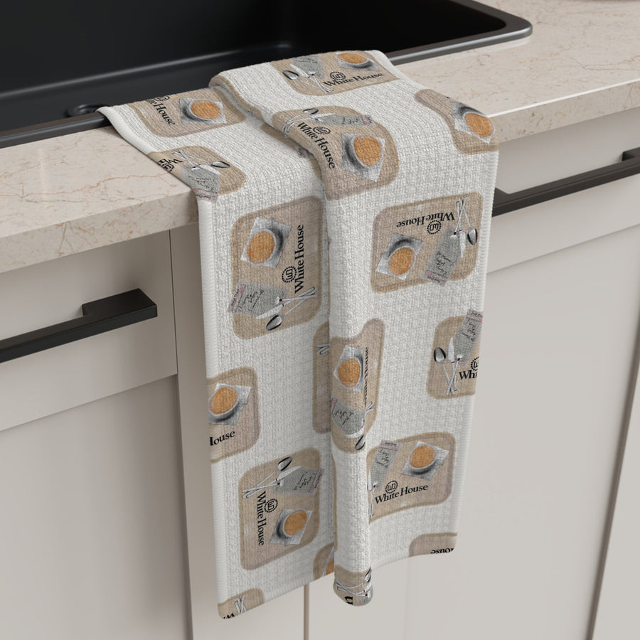 White House Cafeteria tea towel draped beside a black kitchen sink in a modern cream kitchen. The light oatmeal towel features a repeating pattern of vintage cafeteria trays with coffee, silverware, a guest check and the White House name from Bismarck, North Dakota.