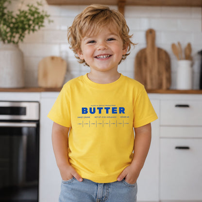 Little boy wearing yellow butter toddler shirt with retro foodie bakery graphic in modern kitchen setting. Boutique kids t-shirt designed for preschool outfits, birthdays, baking families, and playful everyday wear.