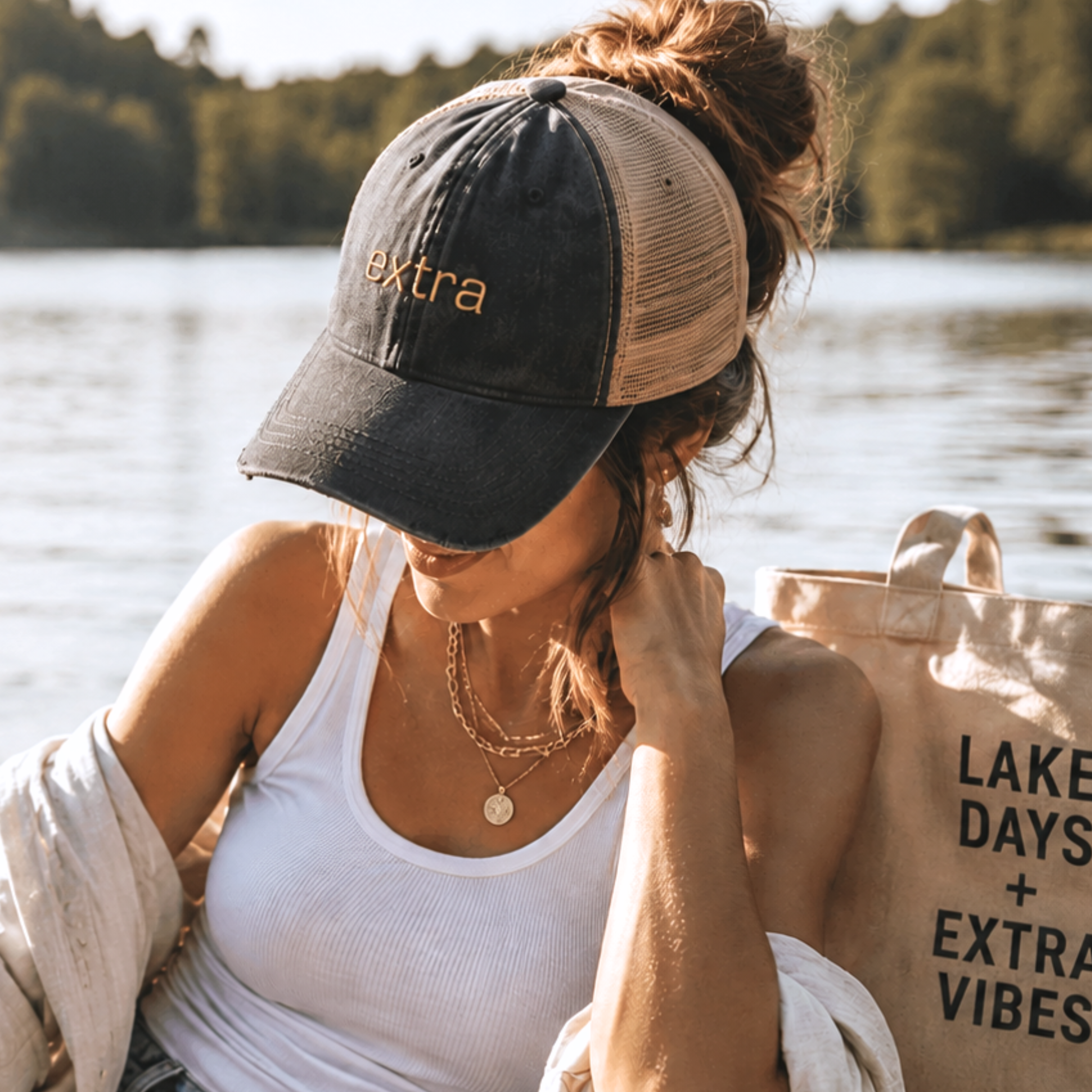 Woman wearing black embroidered extra trucker hat with messy bun beside tote bag at a sunny lakeside outdoor setting.