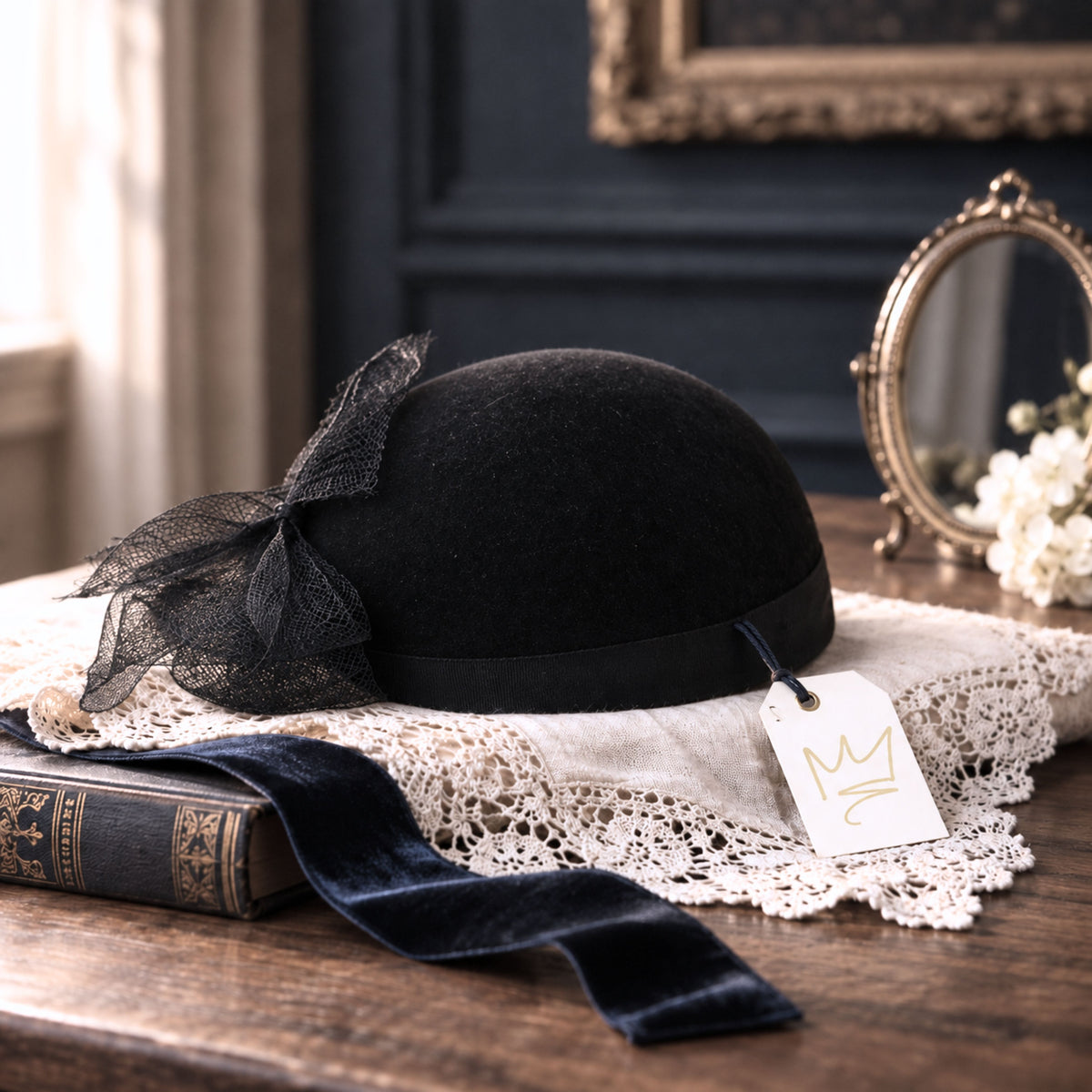 Vintage black felt hat with net trim styled on ivory lace beside a navy velvet ribbon, antique book, brass mirror and gold crown tag.