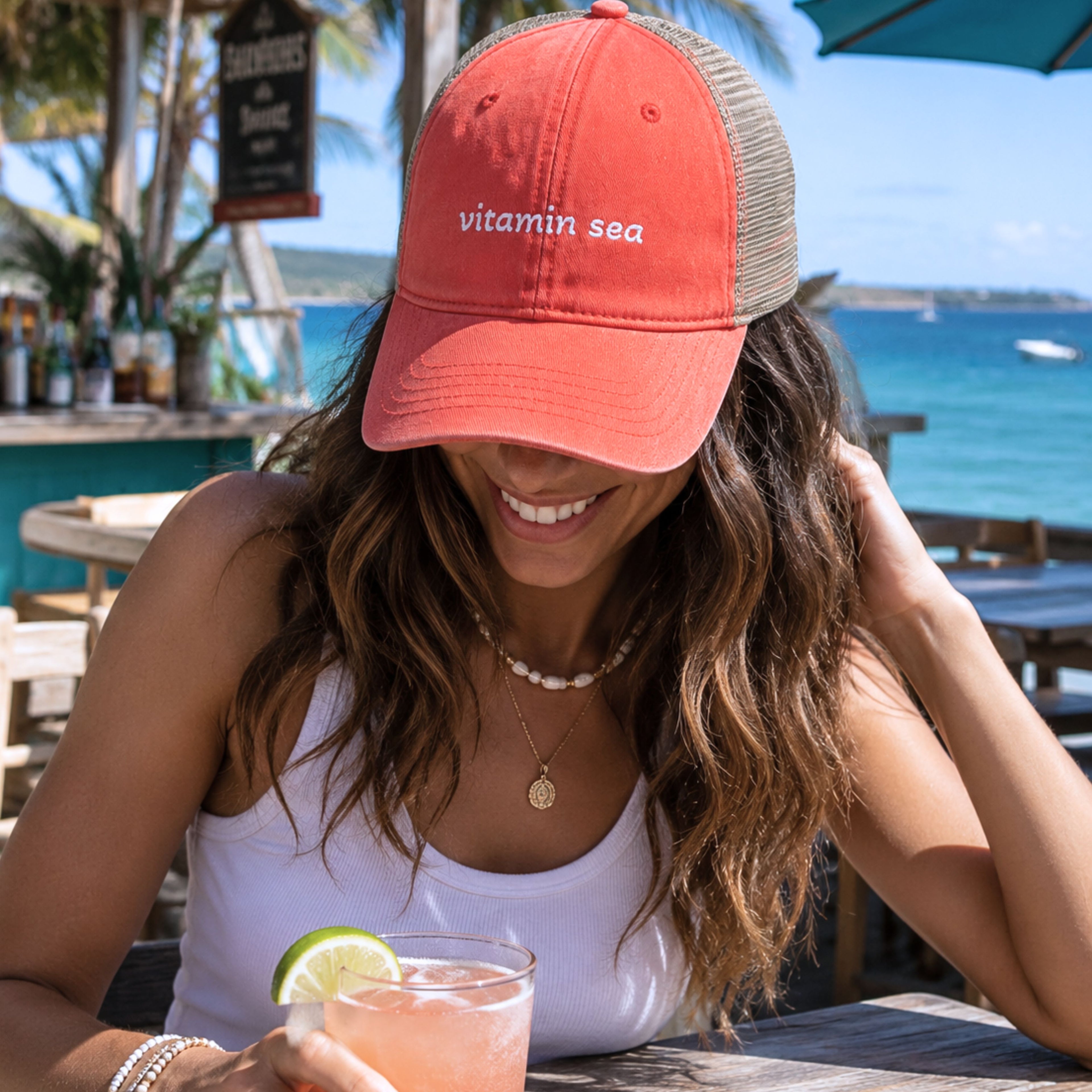 Woman wearing vitamin sea coral trucker hat at a tropical seaside patio bar with ocean views