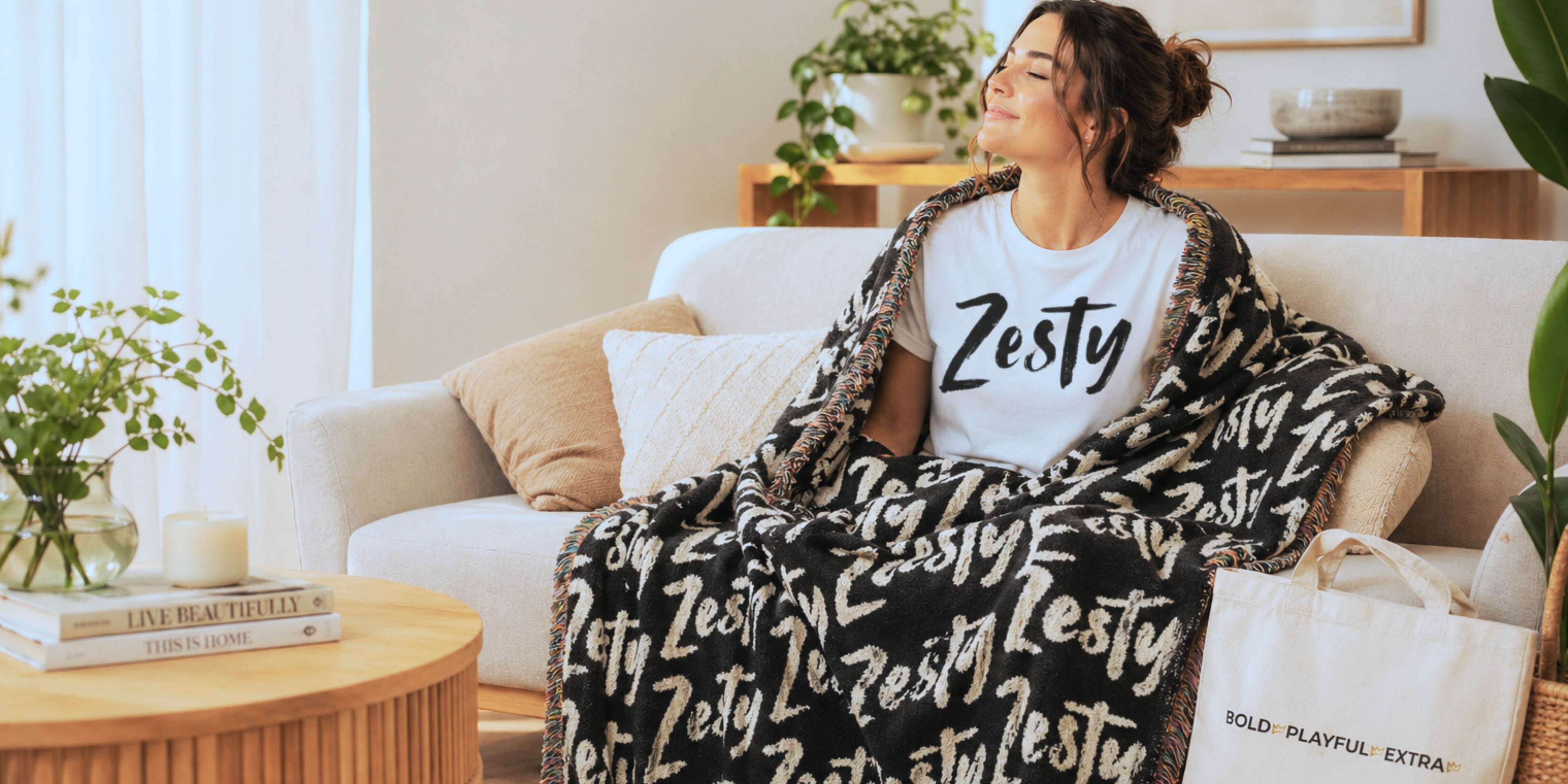 Bright cozy living room lifestyle banner with woman wearing Zesty tee, wrapped in Zesty blanket, styled with canvas tote and warm neutral modern decor for relaxed weekend vibe