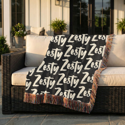 Black blanket with 'Zesty' text draped over a wicker chair outdoors.