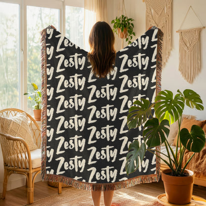 Person holding a black blanket with 'Zesty' text in a cozy room with plants and decor.
